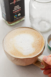 A latte made without a machine on a white countertop with a hand holding it.