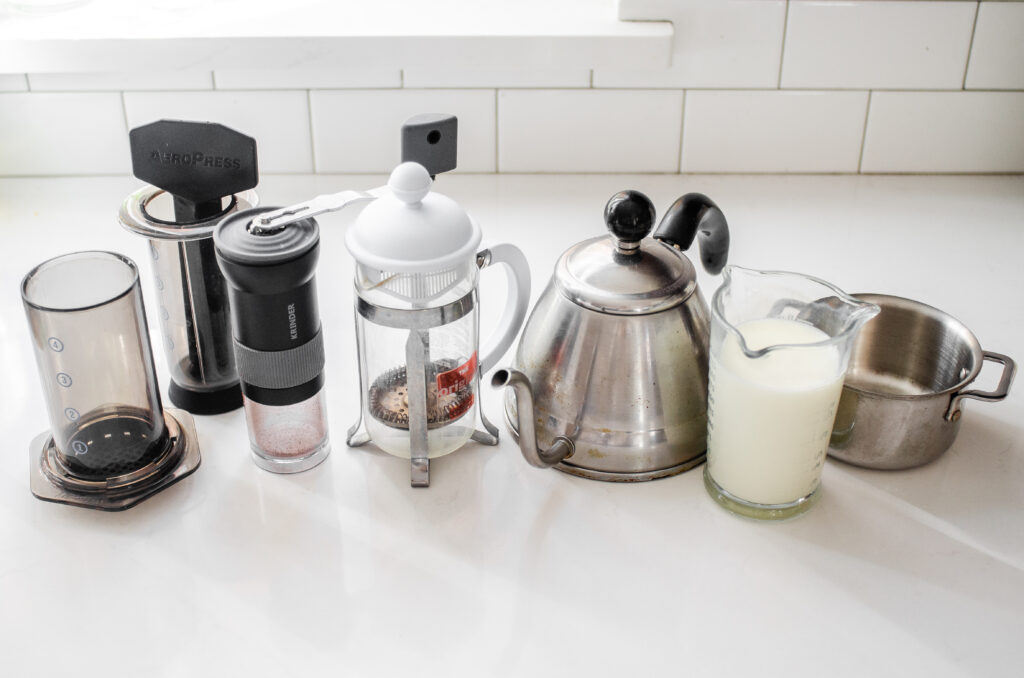 An aeropress, hand coffee grinder, French press, tea kettle, measuring cup of milk, and small pot on a white countertop.