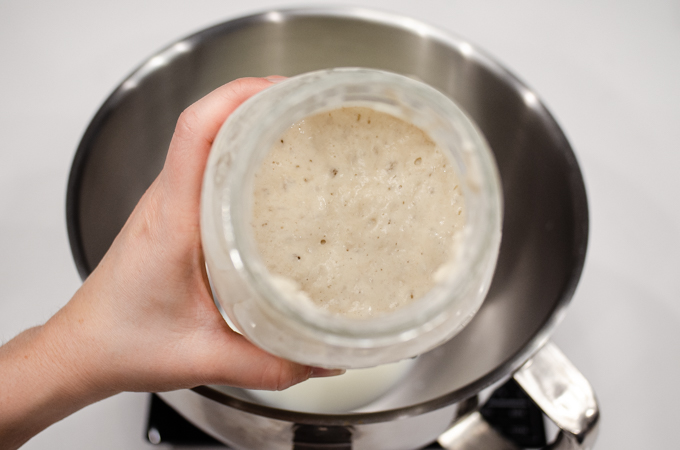 A jar of sourdough starter being held over the bowl of a stand mixer.