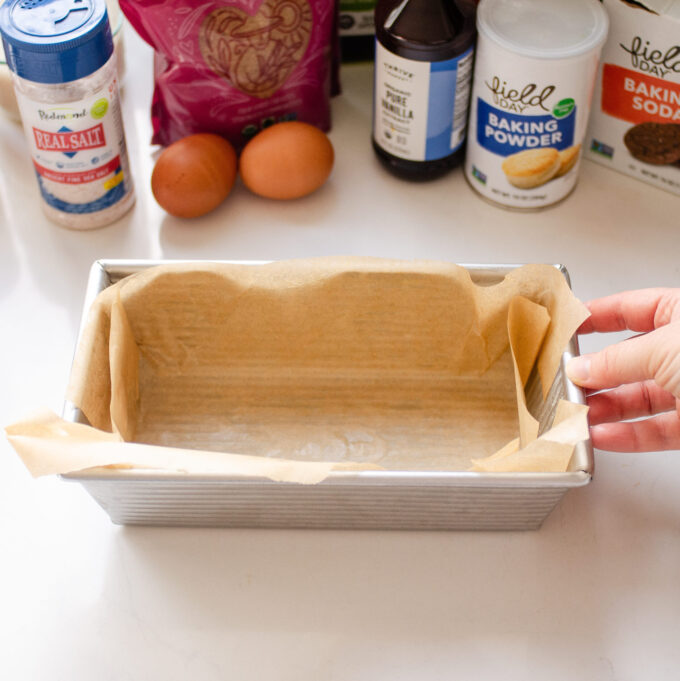 A loaf pan neatly lined with parchment paper with a hand holding one edge. 
