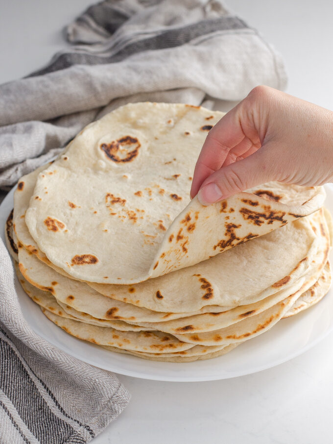 Sourdough tortillas stacked on a plate with a tea towel around them with a hand rolling the top tortilla.