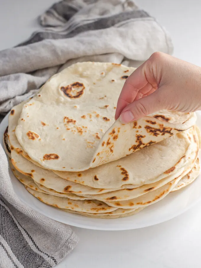 Sourdough tortillas stacked on a plate with a tea towel around them with a hand rolling the top tortilla.