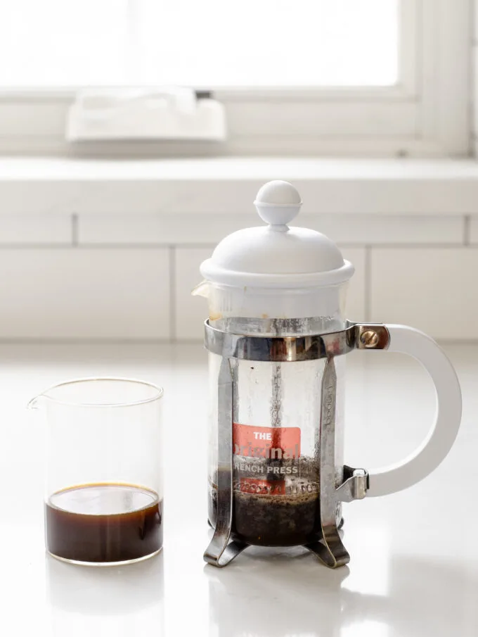 2 shots of espresso strength coffee in a clear pitcher next to a French press on a white countertop.