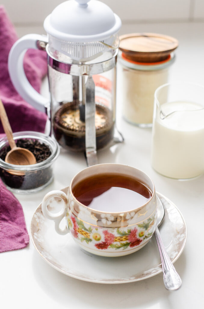 A teacup filled with black tea with a French press, sugar, and cream behind it.