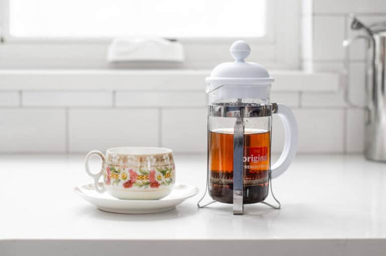 A teacup with a French press filled with black tea next to it.