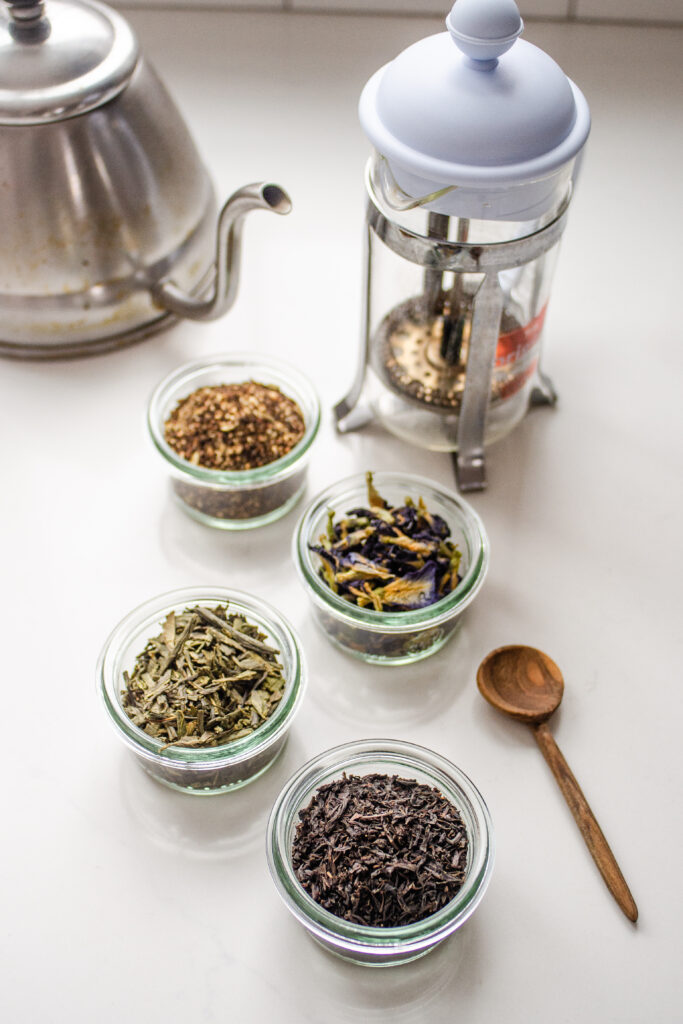 An assortment of different types of tea on a white countertop with a tea kettle on one side, and a French press in the back.