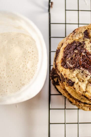 A stack of chocolate chip sourdough cookies with a jar of sourdough starter discard to the side.