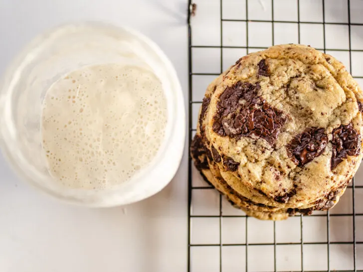 A stack of chocolate chip sourdough cookies with a jar of sourdough starter discard to the side.