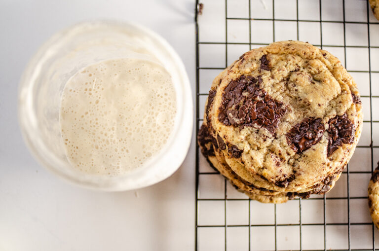 A stack of chocolate chip sourdough cookies with a jar of sourdough starter discard to the side.