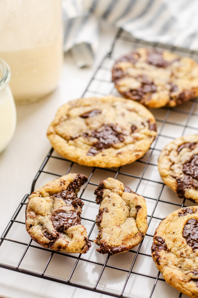 A broken sourdough chocolate chip cookie on a wire cooling rack with other cookies around it and a glass of milk off to the side and a jar of sourdough discard.