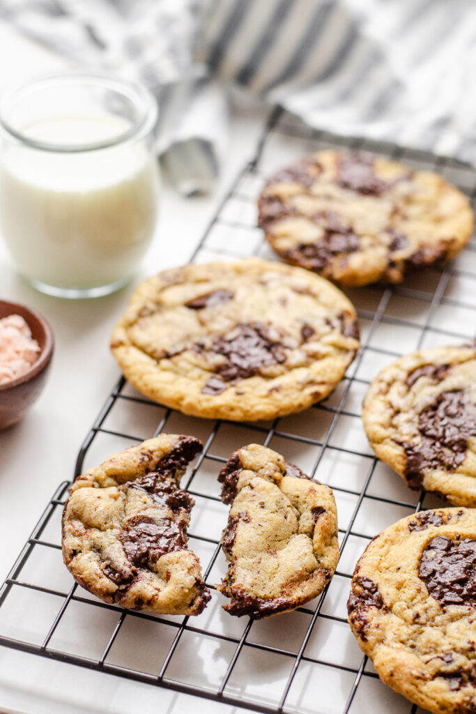 A broken sourdough chocolate chip cookie on a wire cooling rack with other cookies around it and a glass of milk off to the side.