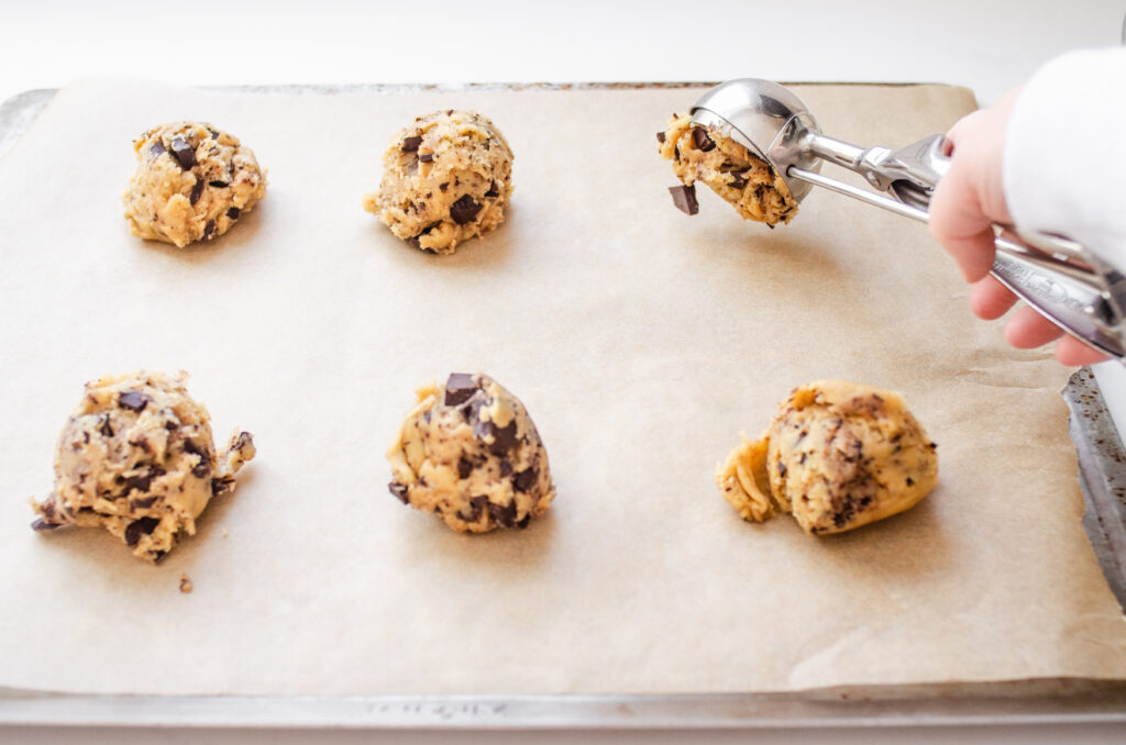 Scooping chocolate chip cookie dough onto a parchment lined baking sheet with a cookie scoop.