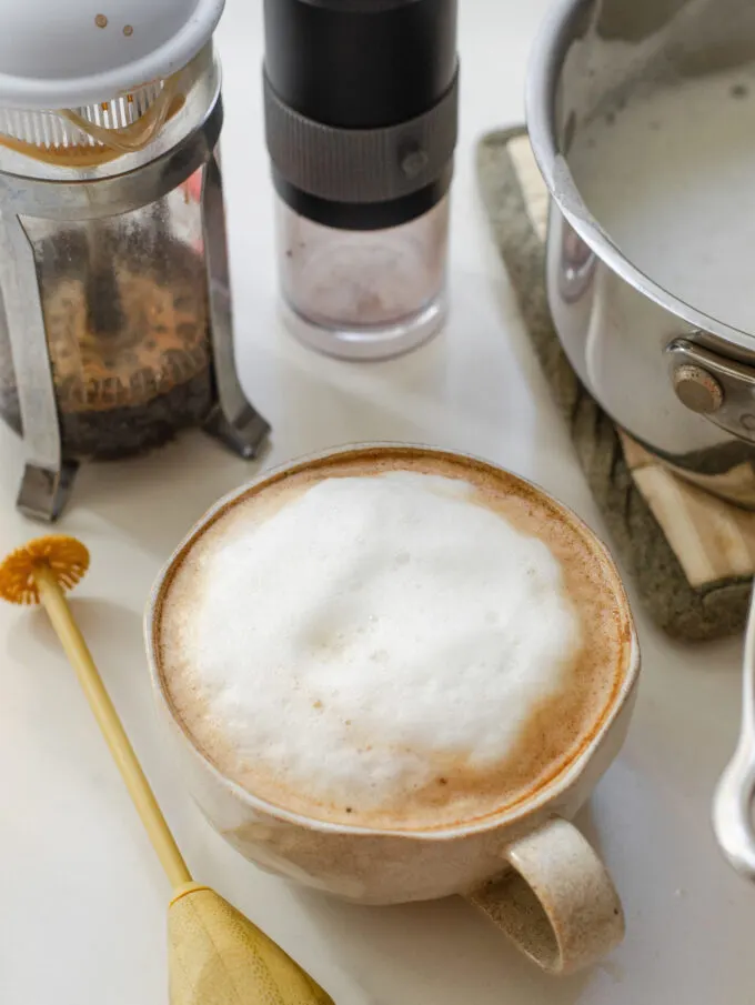 A latte made with a French press and hand held frother on a white countertop with the French press behind the latte mug.