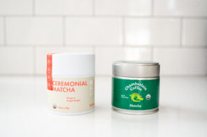A tin of Rishi matcha next to a tin of Chamberlain matcha on a white countertop with a subway tile backsplash. 