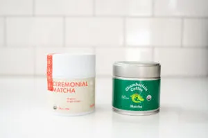 A tin of Rishi matcha next to a tin of Chamberlain matcha on a white countertop with a subway tile backsplash.
