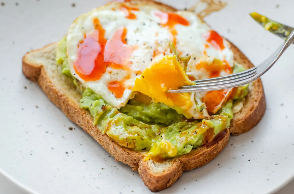 A fried egg (over medium) on avocado toast with a piece of the yolk on a fork.