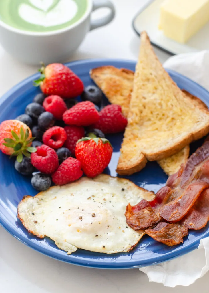 A fried over medium egg on a blue plate with bacon, fresh berries, and toast triangles around it.