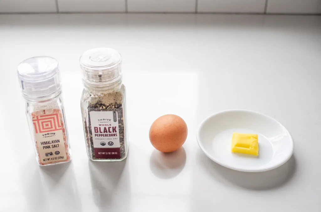 Salt and pepper shakers, a brown egg, and a small pat of butter laid out on a white countertop.