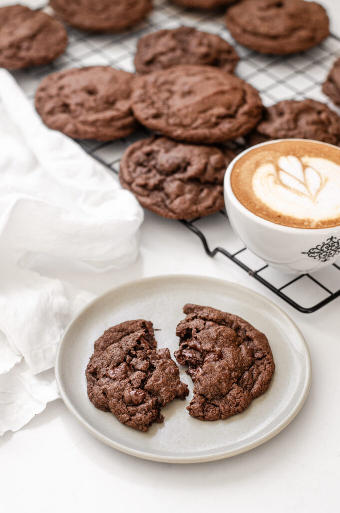 A chocolate espresoo cookie on a plate with a latte and more cookies in the background on a wire rack.