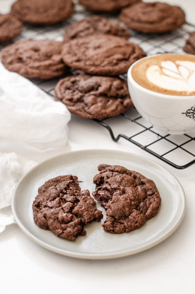 A chocolate espresoo cookie on a plate with a latte and more cookies in the background on a wire rack.