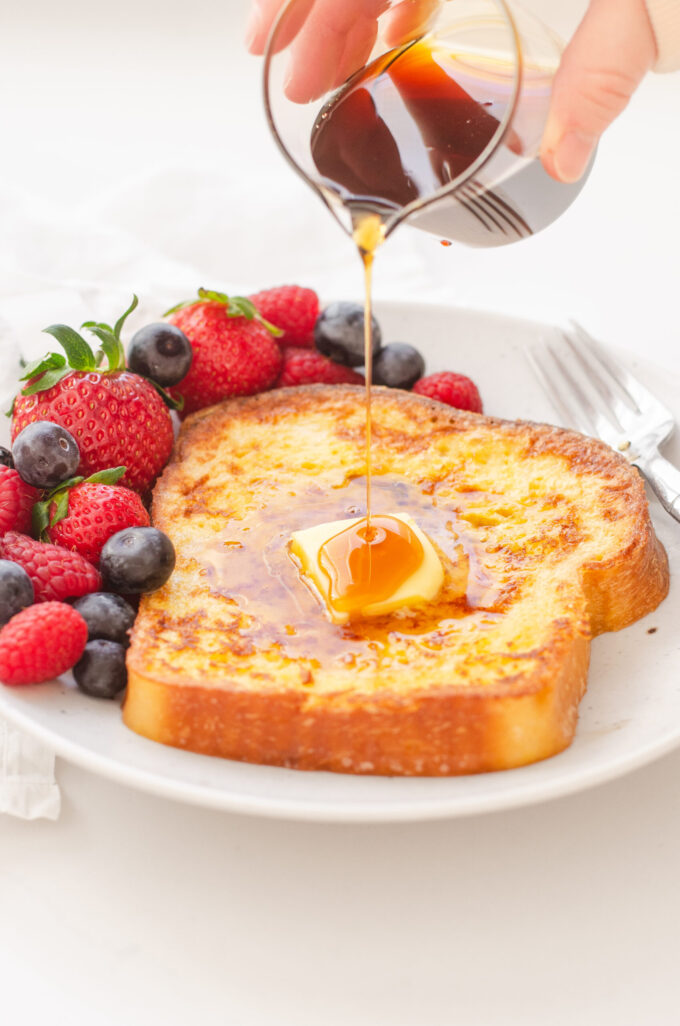 Pouring maple syrup onto a buttered piece of French toast with fresh berries around it.