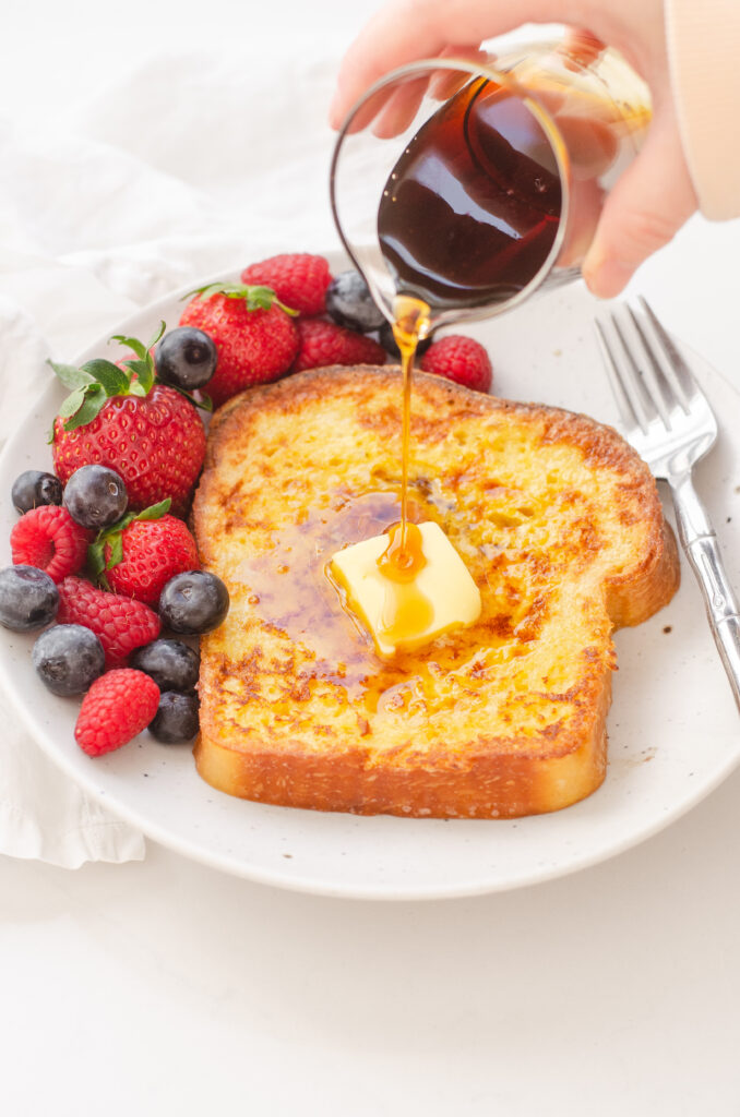 Pouring maple syrup onto a buttered piece of French toast with fresh berries around it.
