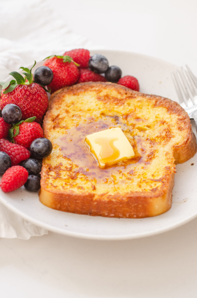 A piece of French toast on a plate with fresh berries off to one side, and a fork on the other.