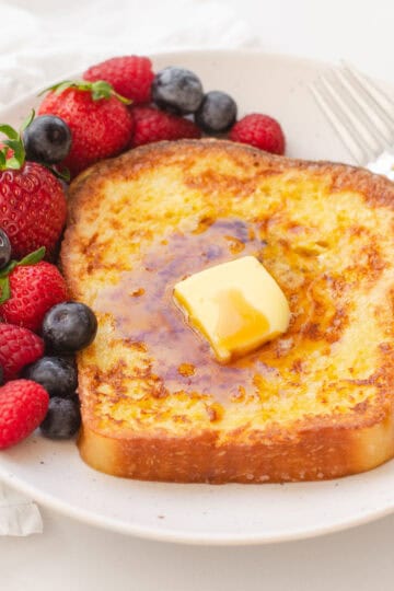 A piece of French toast on a plate with fresh berries off to one side, and a fork on the other.