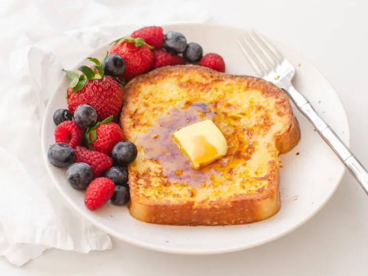 A piece of French toast on a plate with fresh berries off to one side, and a fork on the other.