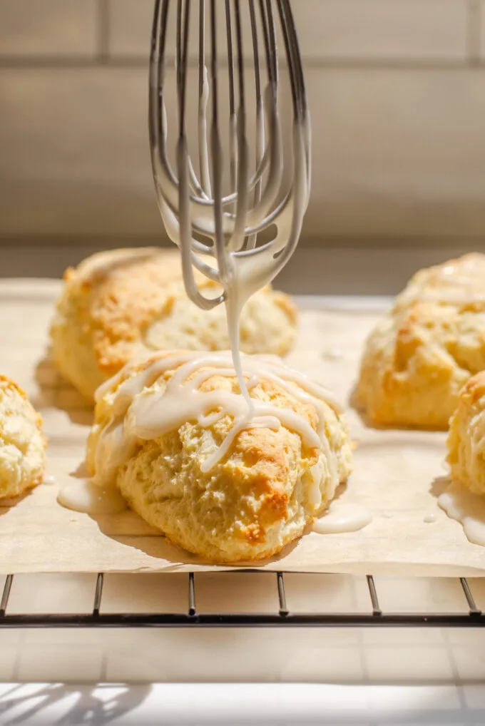 Drizzling a glaze on top of lemon scones on parchment paper on top of a wire cooling rack.