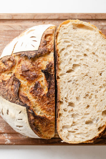 A top down shot of a loaf of sourdough bread that's been baked in a Dutch oven with half upright, and the other half showing the crumb shot.