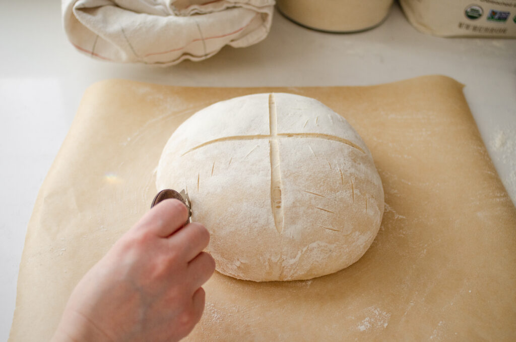Scoring a round loaf of sourdough bread with an X pattern.