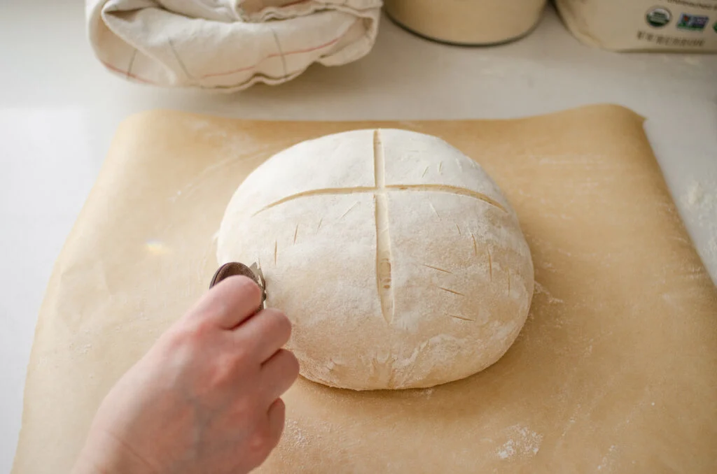 Scoring a round loaf of sourdough bread with an X pattern.
