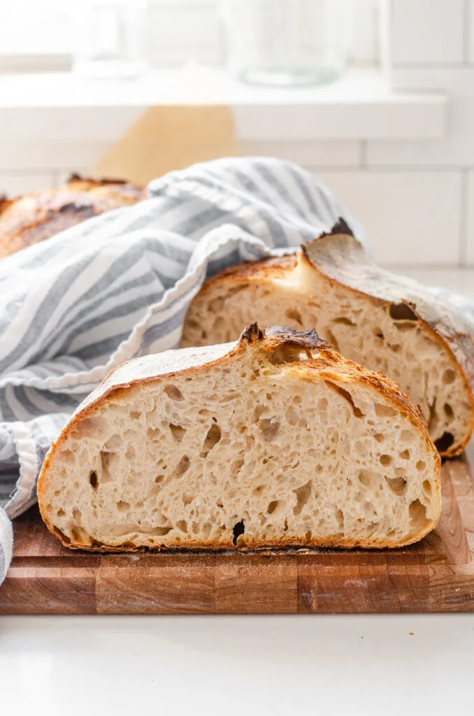 A loaf of sourdough bread cut in half on a wooden cutting board with the other half and a striped linen tea towel behind it.