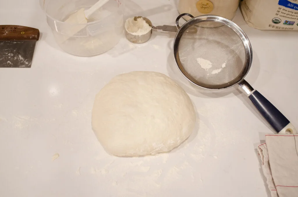 A ball of sourdough bread dough on a white countertop with a fine mesh sieve next to it.