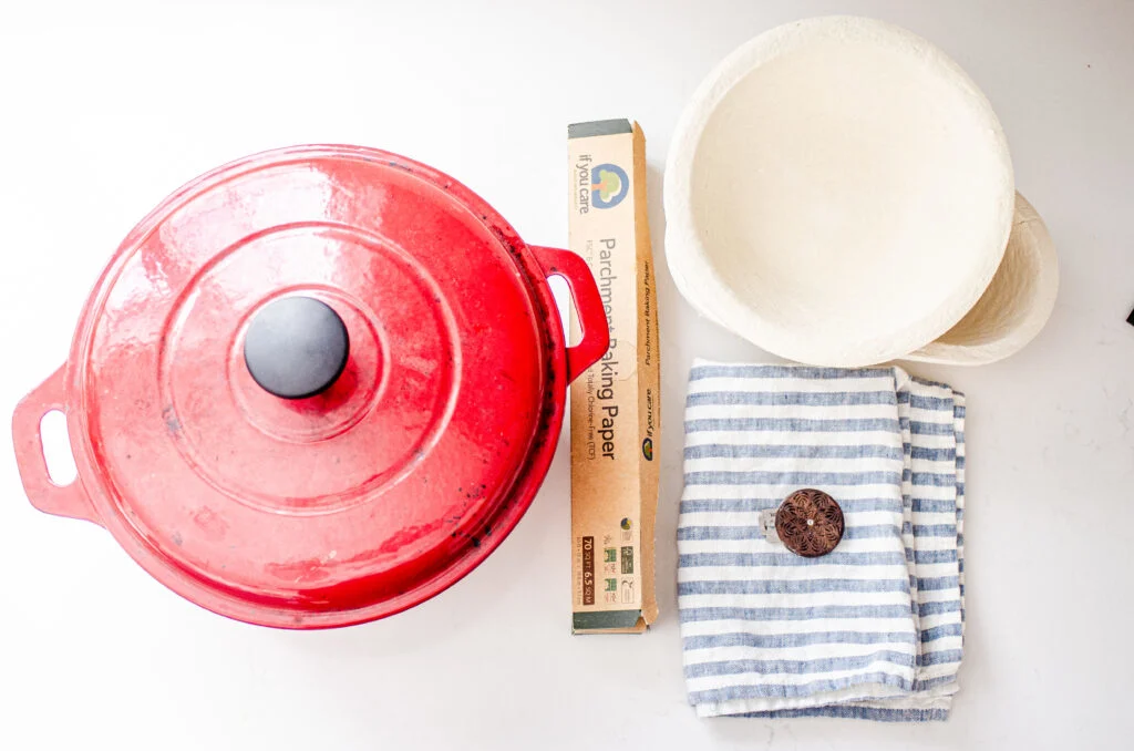 A red Dutch oven, a roll of parchment paper, banneton baskets, a tea towel, and a lame.