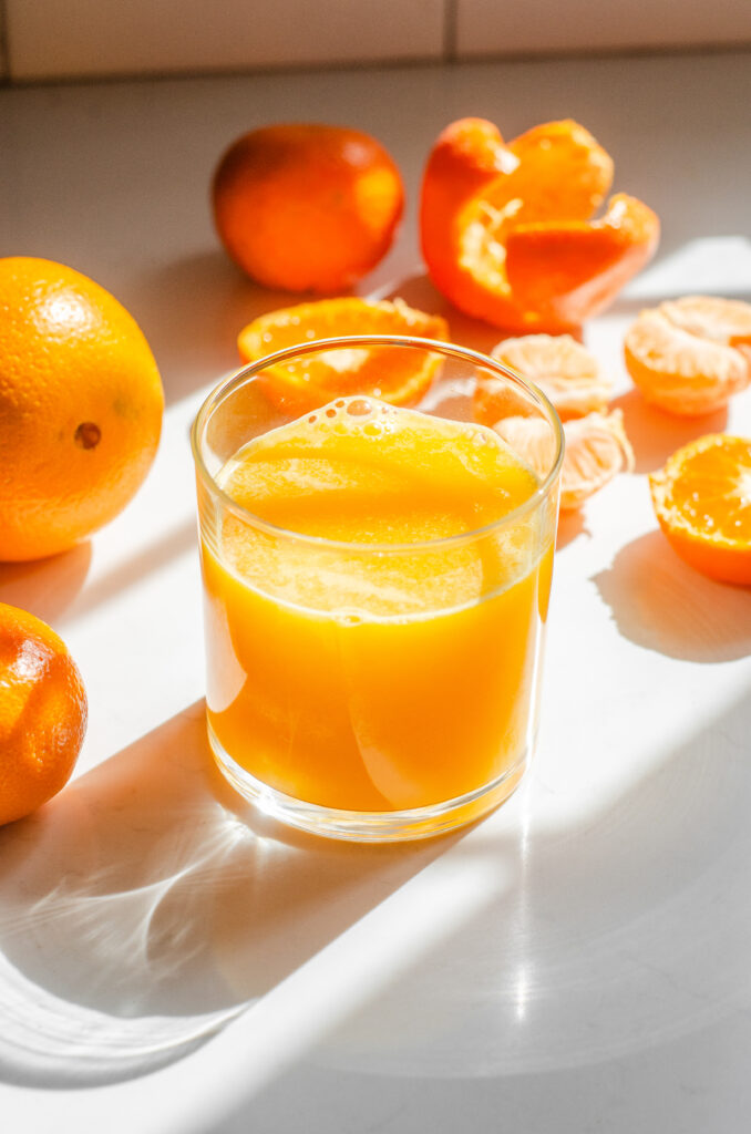 A glass of freshly squeezed orange juice on a sunny countertop with oranges in various states of being processed around it.