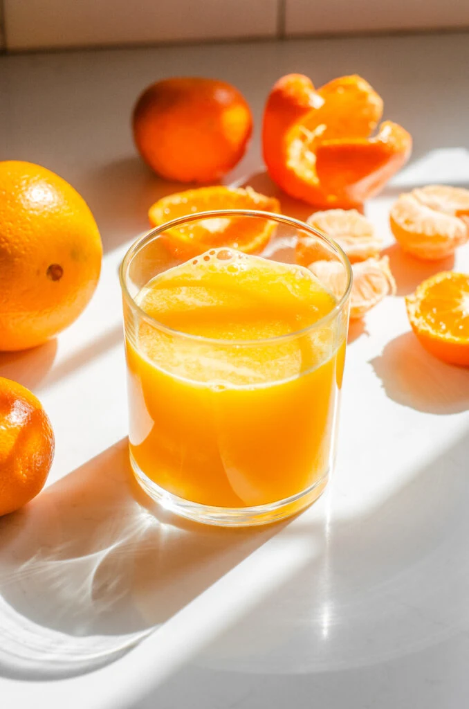 A glass of freshly squeezed orange juice on a sunny countertop with oranges in various states of being processed around it.