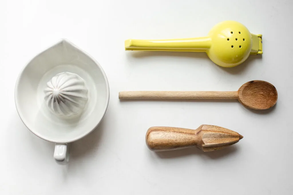 A citrus juicer, citrus press, wooden spoon, and citrus reamer laid out on a white countertop.