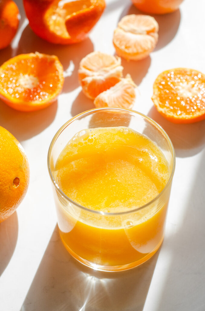 A glass of freshly squeezed orange juice on a sunny countertop with oranges in various states of being processed around it.
