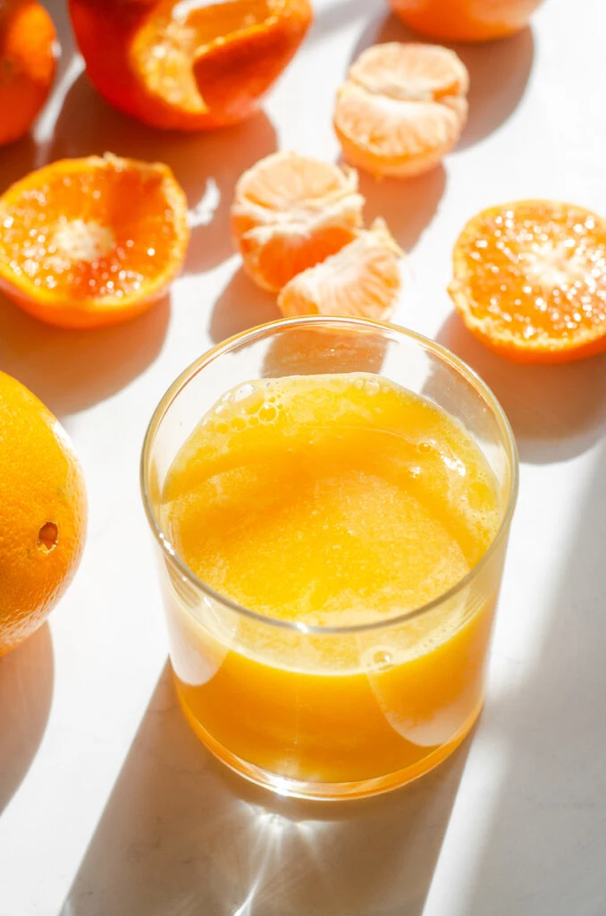 A glass of freshly squeezed orange juice on a sunny countertop with oranges in various states of being processed around it.
