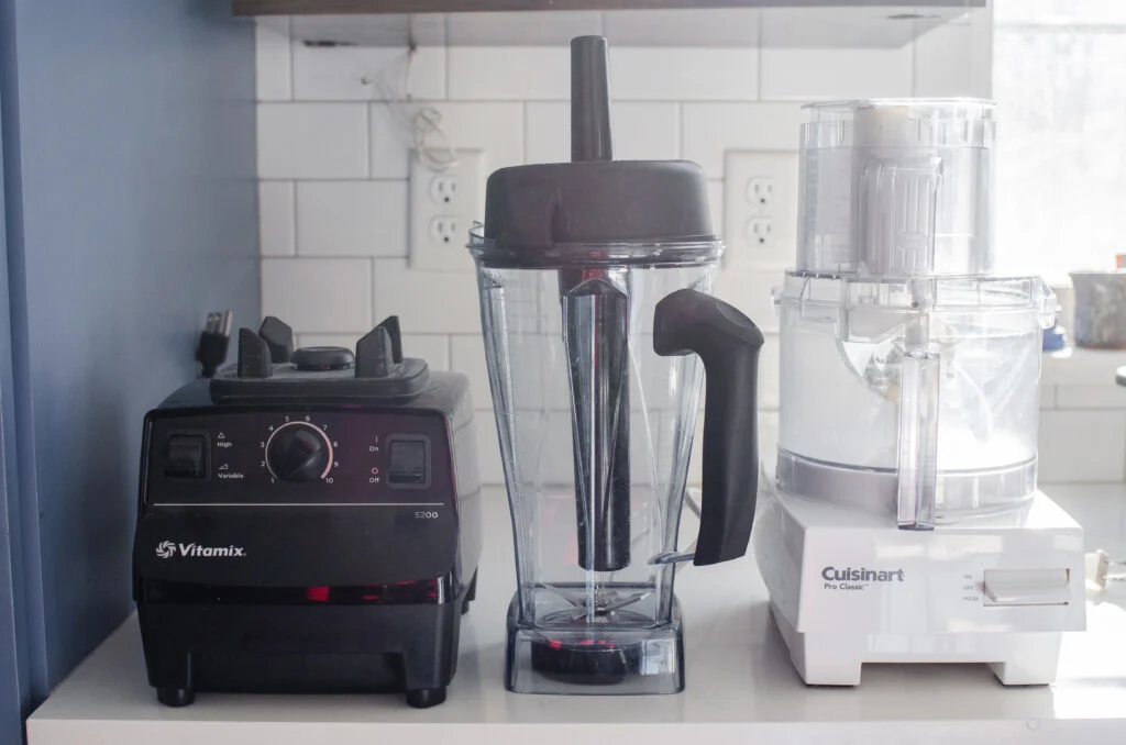 A blender and food processor on a white countertop with a subway tile backsplash behind it.