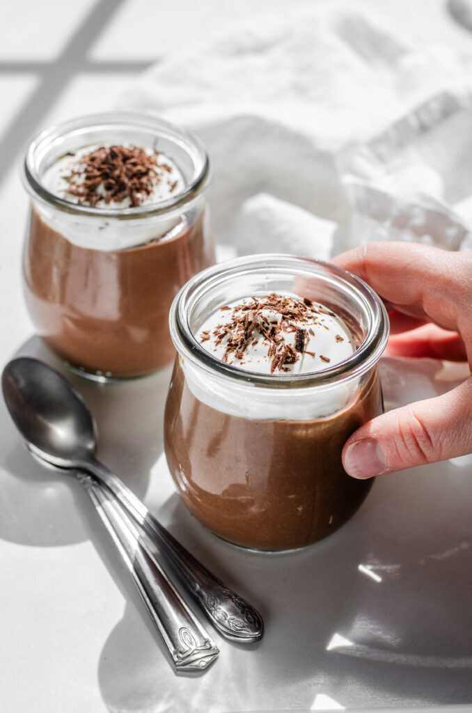 Two jars of homemade chocolate pudding with whipped cream and chocolate shavings on a white countertop with a napkin in the background and a hand holding one of the jars.