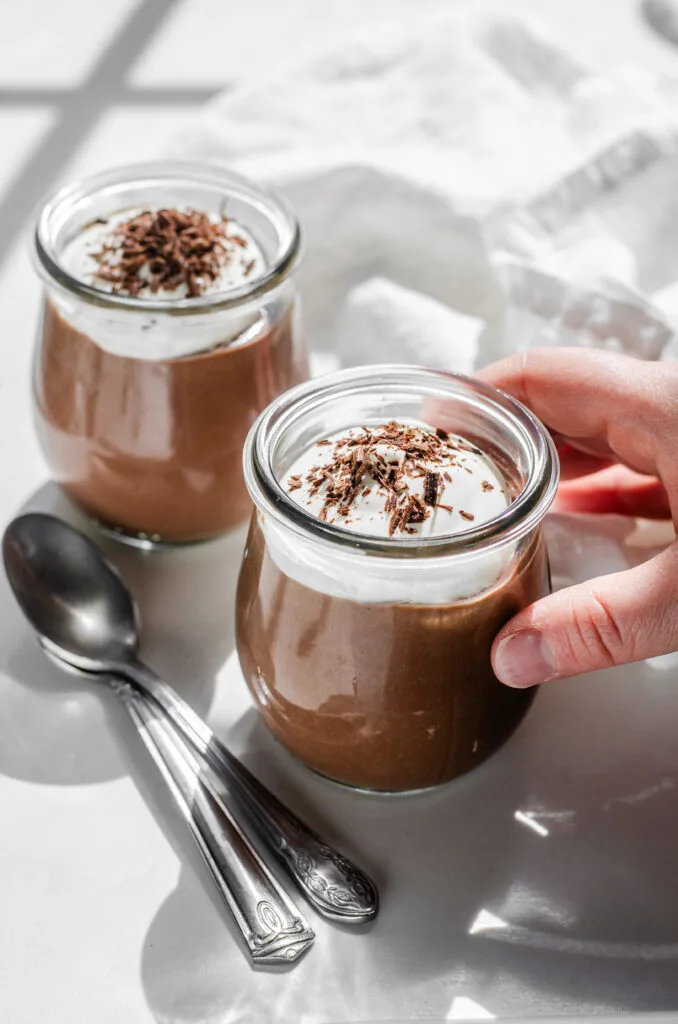 Two jars of homemade chocolate pudding with whipped cream and chocolate shavings on a white countertop with a napkin in the background and a hand holding one of the jars.