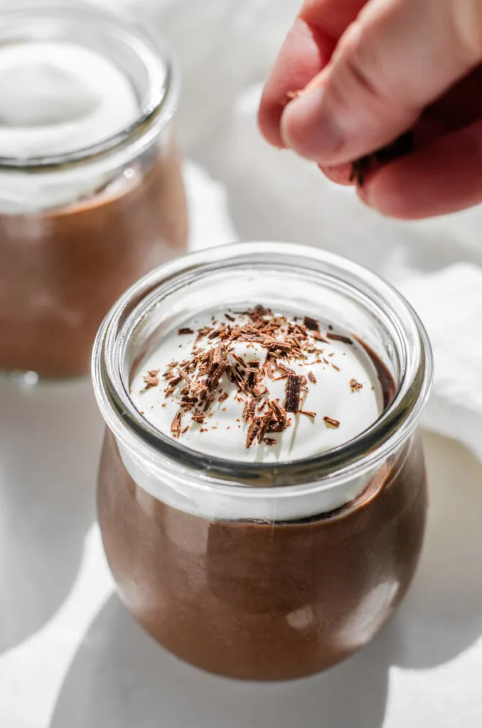 A hand sprinkling chocolate shavings on top of a jar of chocolate pudding topped with whipped cream.