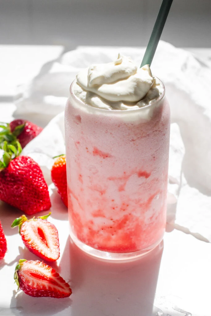 A homemade strawberry frappuccino on a white countertop with fresh strawberries on one side, and a linen napkin on the other.