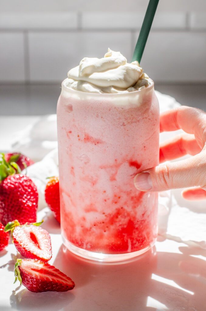 A 16 ounce glass filled with a strawberry frappuccino with whipped cream on top, fresh strawberries off to the side, and a hand holding the cup.