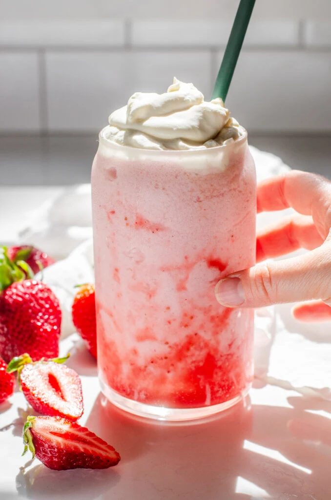 A 16 ounce glass filled with a strawberry frappuccino with whipped cream on top, fresh strawberries off to the side, and a hand holding the cup.