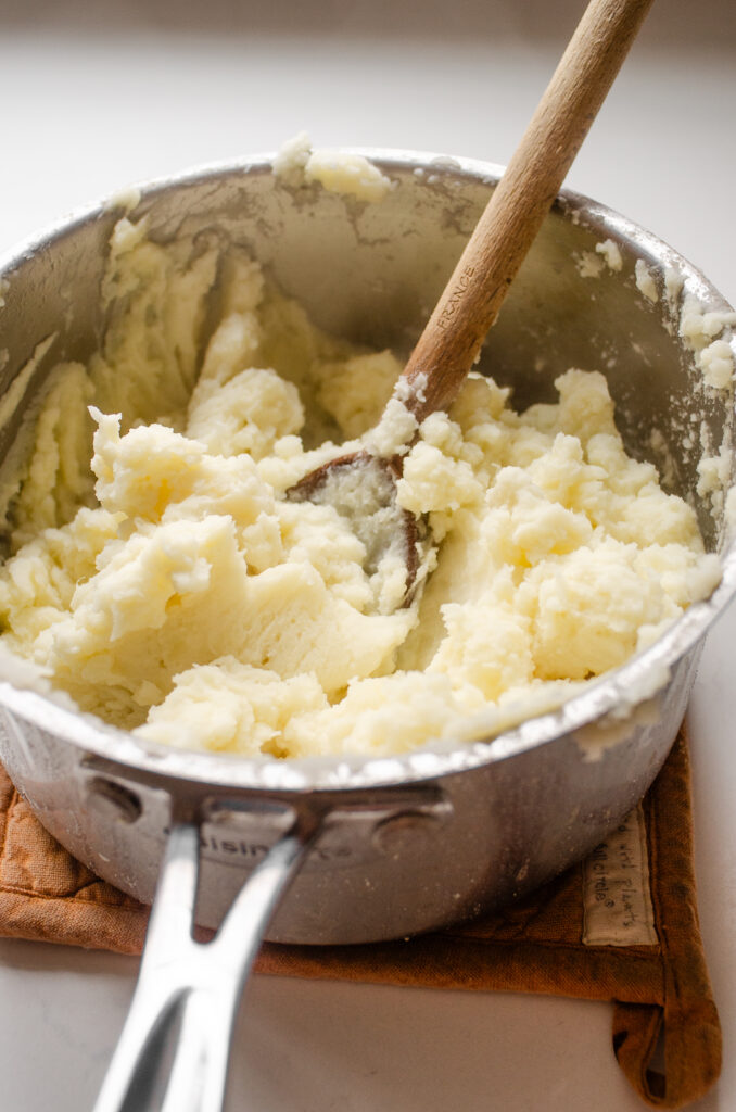A pot filled with mashed potatoes with a wooden spoon inside to show how they were mashed.