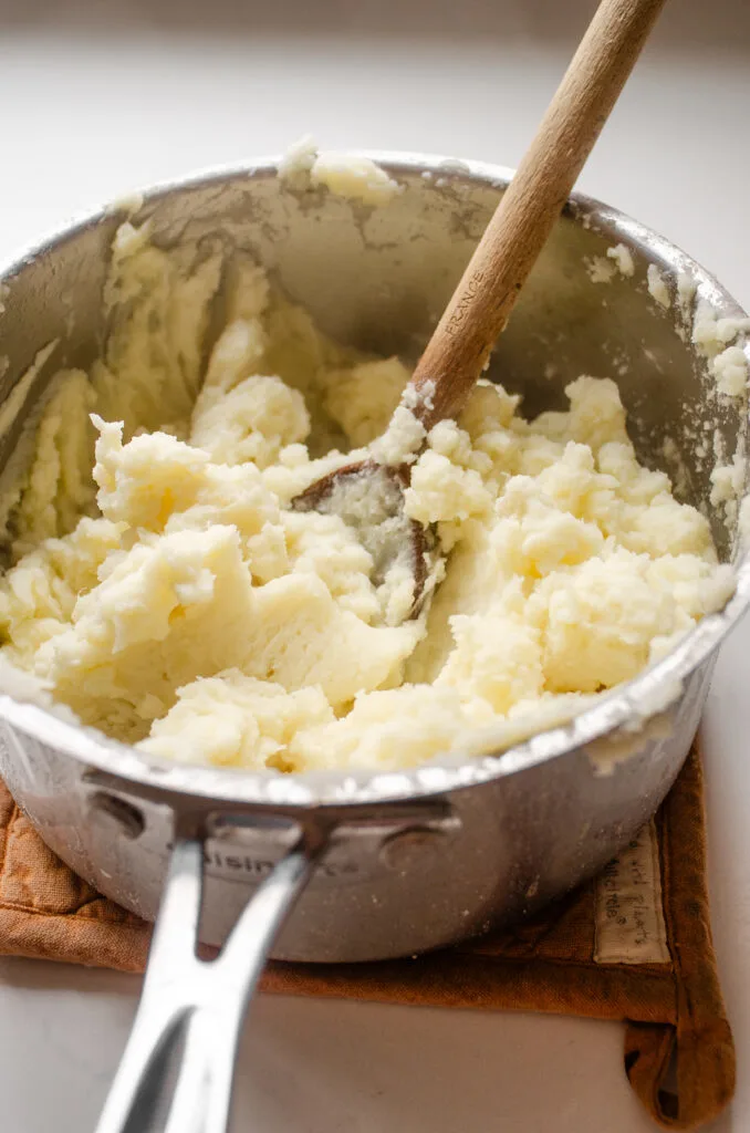 A pot filled with mashed potatoes with a wooden spoon inside to show how they were mashed.